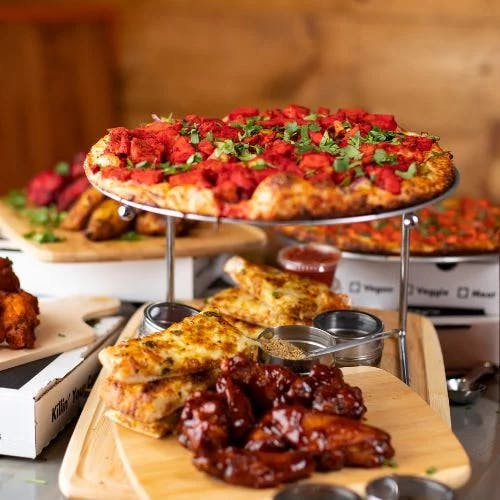 Clickable image showing A crisp hot pizza topped with chicken and fresh herbs, with a side of bread and barbequed wings. Should open an Instagram external link to the source image.